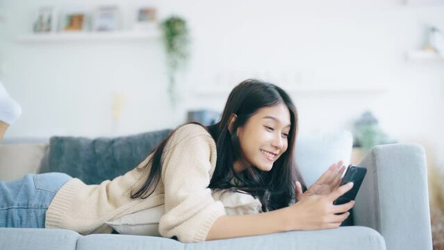 Happy Young Asian Woman Relaxing at Home. Smiling on Sofa with Mobile Smartphone. Video Call with Friend