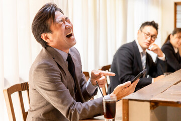 カフェの店内でスマホを見て大声で笑う迷惑なビジネスマンの男性客
