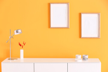 Candles, lamp on chest of drawers and blank frames hanging on orange wall in room