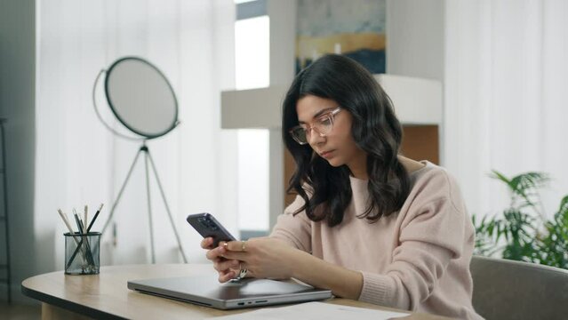 Business people concept. Pensive hispanic marketing manager in stylish eyewear looking at smartphone. Beautiful smart woman using mobile phone sitting at desktop remote home office in modern apartment