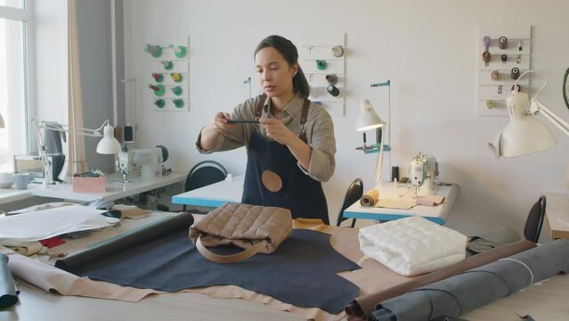 Full shot of young biracial craftswoman standing in leather workshop and taking photos of new handbag design on smartphone for advertising on social media