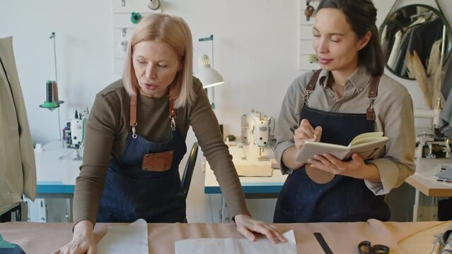 Medium tilting shot of middle-aged Caucasian female fashion designer teaching apprentice in leather workshop, and young woman listening, asking questions and writing in notebookMedium tilting shot of 