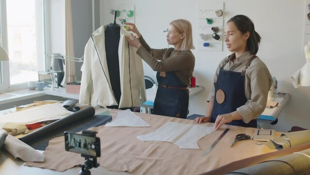 Full arc of two Caucasian female seamstresses standing in front of smartphone on tripod and cutting table in leather workshop, and demonstrating jacket on tailors dummy while recording sewing tutorial