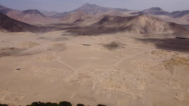Aerial footage of the ancient city of Caral. Drone hovers above and pans slowly pans left, revealing the many desert mountains and pyramids and river and green valley of the Supe Valley in Lima, Peru.