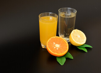 Two glasses with different citrus juices on a black background, next to half a lemon and an orange with leaves.