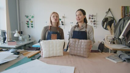 Full shot of two female leather workshop owners standing at table with new handmade tote bags and presenting features and functions while recording story or reel for blog on camera