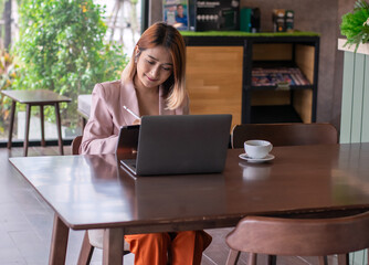 Businesswoman wearing a suit pink sitting at the coffee shop and using laptop conference video call to talk online meeting and using tablet pencil to write report on accounting finances business.
