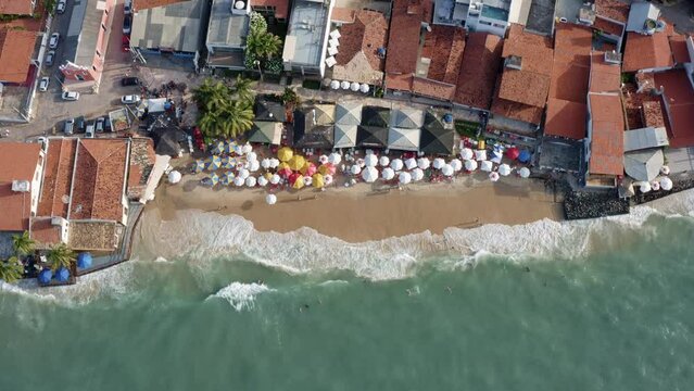 Aerial drone birds eye shot of the beautiful tropical famous Pipa beach during high tide with tourists playing in the water and enjoying the shade under colorful umbrellas during a warm summer evening