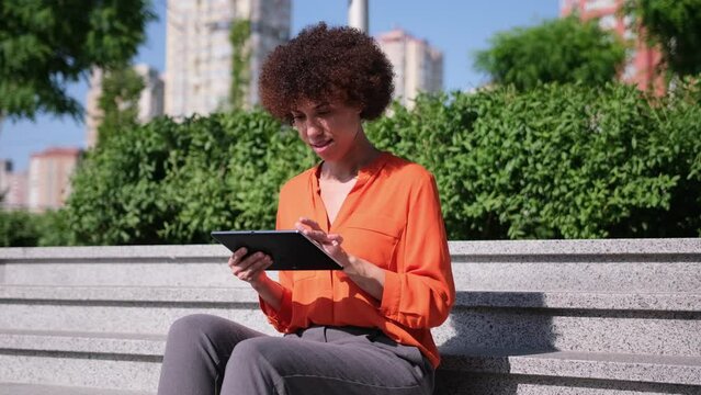 Happy young African American woman using digital tablet pad sufring internet inspired with new ideas sitting outdoors