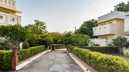 The concrete road in the park is fenced with hedges. Decorative lamps are installed in the trimmed bushes. Flowering trees, palms on lawns. Cottages against the blue sky. India.