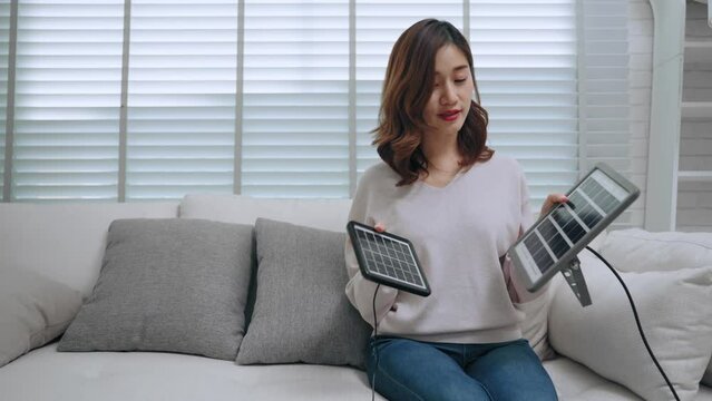 Happy Asian woman sitting on sofa holding two panel of solar cell comparison in living room at home. Sustainable energy and electricity Saving concept