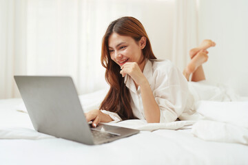 Happy smiling asian woman stretching on bed in the morning and using laptop computer