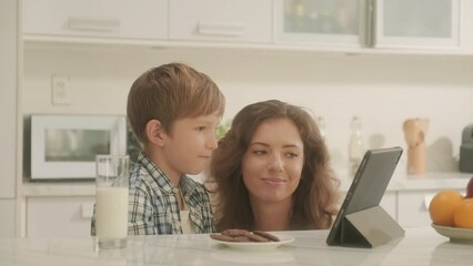 Boy watching video on digital tablet sitting at kitchen table with glass of milk, mom kissing son on cheekBoy watching video on digital tablet sitting at kitchen table with glass of milk, mom kissing  - Powered by Adobe
