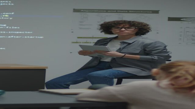 Full shot of female Causian teacher sitting on chair in front of blackboard looking and pointing at screen projection with code explaining it
