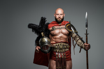Confident bald gladiator with light armor and red cape, holding a spear and gladiator helmet with...