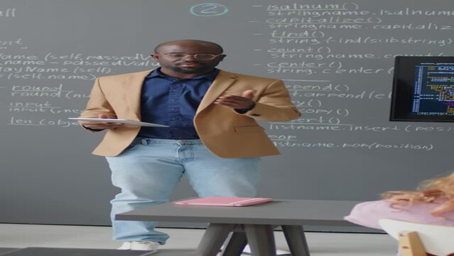 Vertical full shot of male African American teacher giving lecture in front of blackboard showing code on screen in classroom