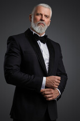 Attractive middle-aged man with grey hair and beard is dressed in a stylish black suit and bowtie,...