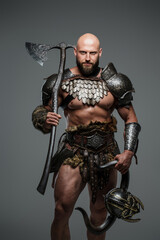 Fototapeta premium A bald bearded viking in lightweight fur-lined armor holding a large two-handed axe stands against a gray background