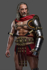 Mature and muscular gladiator with a stylish grey beard and flowing silver locks dons lightweight armor posing against grey backdrop