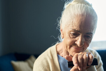 Pensive senior woman with grey hair keeping hand on handle of walking cane while sitting in front...