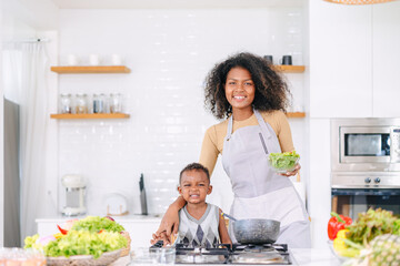 Happy african young mother with her son enjoy eating playful standing together portrait at clean white home kitchen