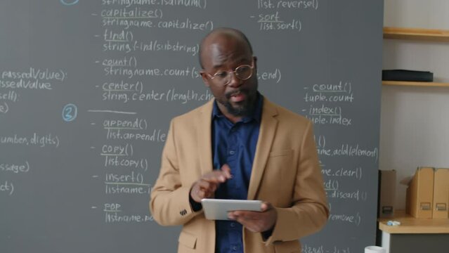 Tracking shot of male African American teacher going there and back in front of blackboard giving lecture on programming in school classroom at daytime