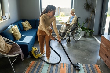 Youthful girl with vacuum cleaner helping her grandmother with disability with domestic chores...