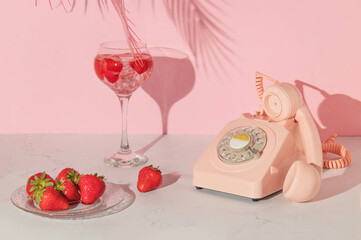 Summer creative layout with cocktail glass, strawberries on plate and retro telephone with pink palm leaf on pastel pink background. 80s or 90s retro aesthetic idea. Minimal summer drink idea.