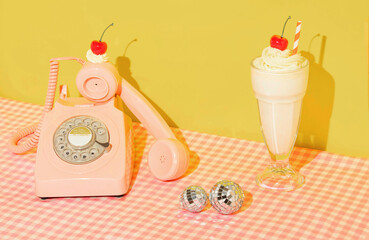 Summer creative layout with pink retro telephone with whipped cream and bright red cherry, milkshake and disco balls on pastel pink plaid and yellow background. 80s or 90s retro aesthetic girl idea. 