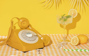Summer creative layout with cocktail glass, lemons, drinking straws, ice cubes and yellow retro telephone with palm leaf on bright yellow and plaid background. 80s or 90s retro aesthetic 