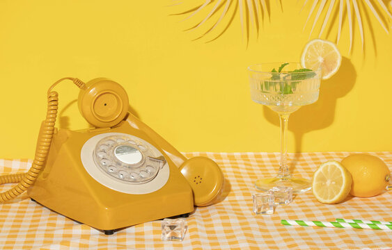 Summer creative layout with cocktail glass, lemons, drinking straws, ice cubes and yellow retro telephone with palm leaf on bright yellow and plaid background. 80s or 90s retro aesthetic  - Powered by Adobe