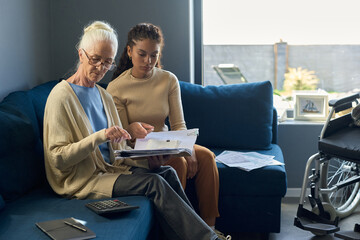 Grandmother and granddaughter pointing at financial bill while sitting on sofa in living room and...