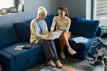 Helpful teenage girl pointing at financial bill while sitting on sofa next to her grandmother and explaining her information in document