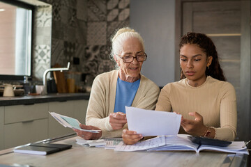 Obraz premium Grandmother and granddaughter sitting by table in the kitchen and discussing financial bills while one of them explaining information