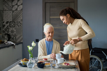 Cute teenage girl with porcelain teapot pouring tea into cup of her grandmother sitting by table in the kitchen during breakfast - Powered by Adobe