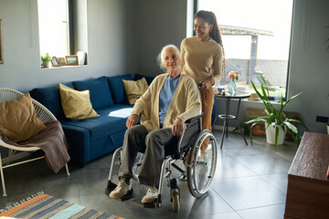 Teenage girl pushing wheelchair with her grandmother while walking along spacious living room with...