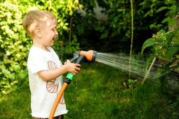 Boy watering flowers and plants in home garden. Child with water hose in summer backyard. Family...