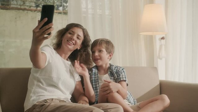 Mother and son in homewear videocalling father via smartphone while sitting on sofa in living room