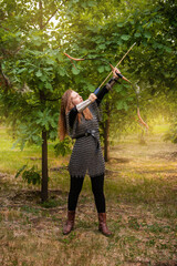 Medieval warrior woman in chain mail and bracers with a bow in her hands against the backdrop of an evening summer forest.