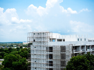 Fototapeta na wymiar A big white concrete construction building is being built surrounded by large green trees on blue sky background with copy space. Modern condominium, apartment or hotel site project.