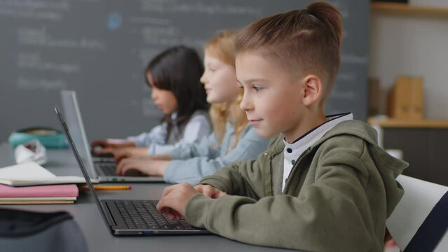 Selective focus on Caucasian elementary age boy sitting at desk doing project work on laptop typing something and two ethnically diverse girls of same age doing same at background