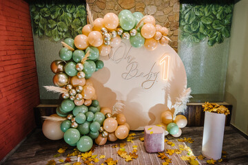 Arch decorated green, brown, golden balloons, text oh baby, neon number one, dry leaves. Photo wall...