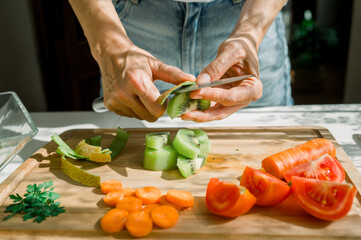 Crop anonymous woman peeling juicy kiwi with knife on wooden cutting board while preparing ingredients for healthy salad in kitchen