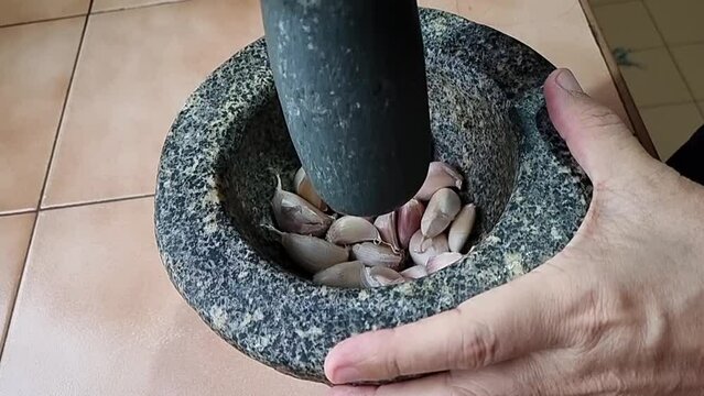 A chef poundes garlic with a pestle in a stone mortar to prepare it for cooking, capturing close-ups in 4K video.