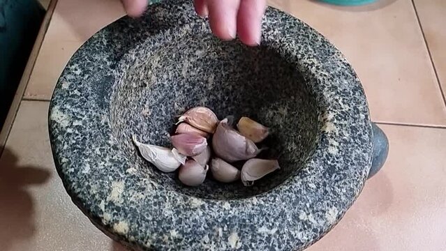 A chef poundes garlic with a pestle in a stone mortar to prepare it for cooking, capturing close-ups in 4K video.