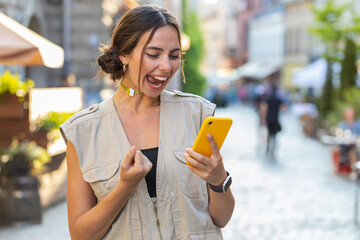 Pretty young woman use mobile smartphone celebrating win good message news, lottery jackpot victory, giveaway online outdoors. Happy girl tourist walking in urban city summer street. Town lifestyles