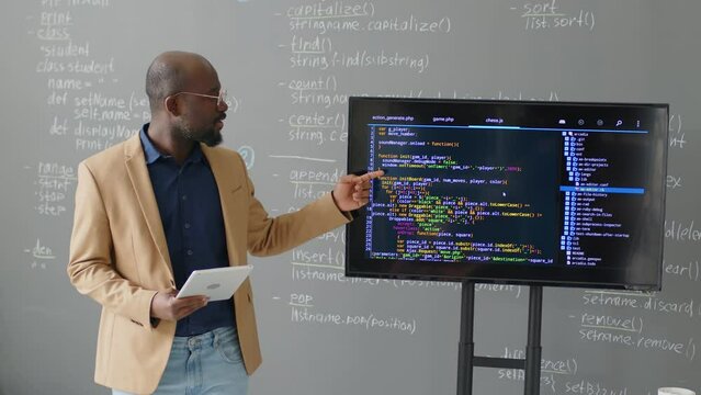 Medium shot of male African American teacher standing in front of blackboard in classroom explaining code on screen at daytime