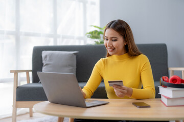 Happy young Asian woman using laptop computer and credit card for online shopping at home. Young Asian female happily utilizes her laptop and credit card for convenient online purchases.