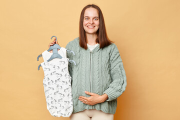 Pleased positive happy young pregnant woman with baby bodysuits isolated over beige background touching her belly looking at camera buying clothing for her future child.