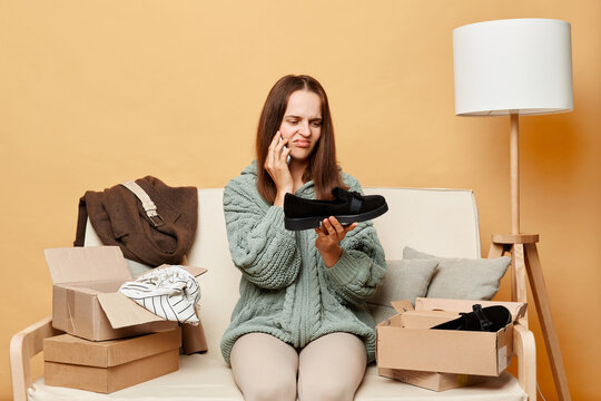 Sad disappointed woman sitting on sofa among boxes with clothing against beige wall talking on smartphone telling about wrong size of her shoes expressing negative emotions.